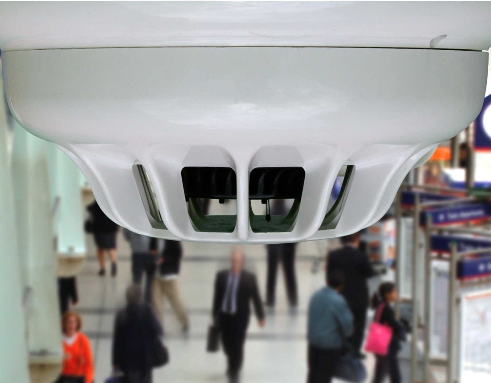Close-up of a fire detection unit mounted on a ceiling, with people walking below in the background